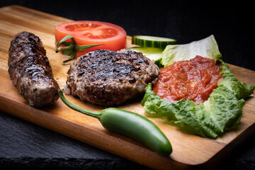 Kyufte, kebapche and lyutenica in a traditional mixed grilled meats dish placed on wooden plank and garnished with green chili pepper, tomato slice and cucumbers.