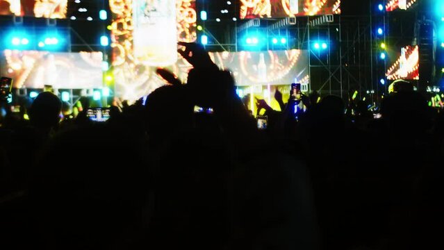 Soft focus Pop band concert on street at night. Silhouettes fans raise their hands in front bright colorful strobe lights on stage.Crowd is dancing, filming performance rock band on their phones.