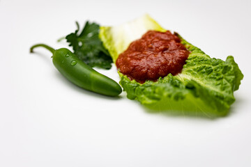Traditional Bulgarian dipping spread sauce called lyutenica made out of tomato paste and ground roasted peppers placed on lettuce leaf next to green chili and parsley isolated on white background