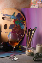 Glass of tasty wine, brushes with colorful paints and burning candle on light gray table