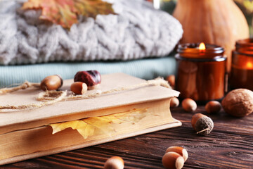 Book with autumn leaf as bookmark, scented candles and warm sweaters on wooden table, closeup