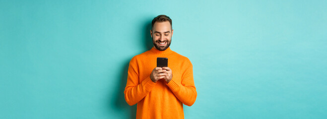Handsome man smiling and texting message on mobile phone, communicating online, standing over light...