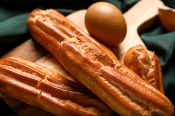 Wooden board with tasty eclairs and egg on dark fabric background, closeup