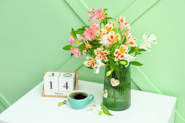 Vase with beautiful alstroemeria flowers, cup of coffee and cube calendar on table near color wall.  Mother's day celebration