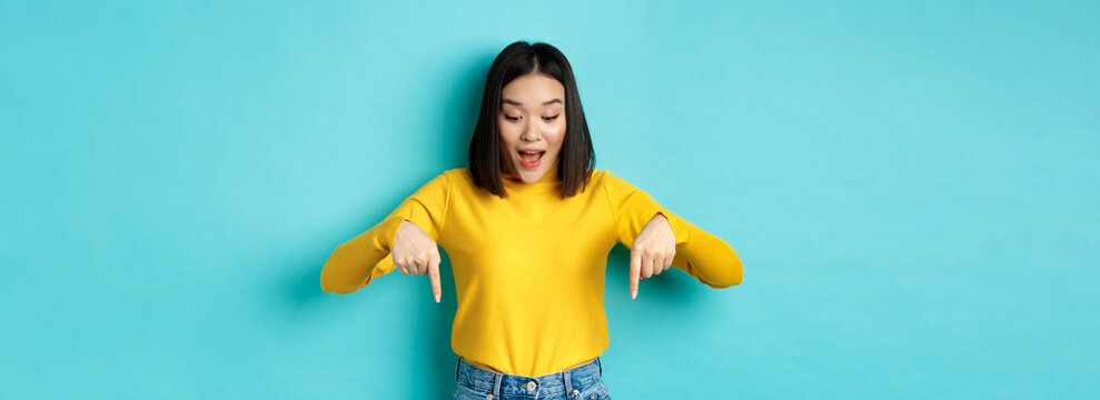 Shopping concept. Surprised cute asian girl checking out discounts, pointing fingers down and looking amazed, saying wow, standing over blue background
