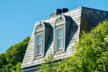 Dual attic windows in the midday sun with pyramid shapped flat roof and front yard trees under clear blue sky