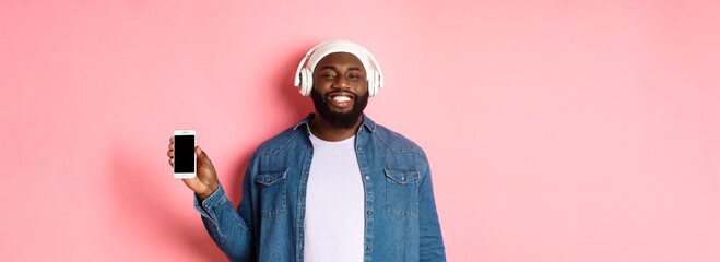 Happy hipster guy listening music on headphones and showing mobile screen, smiling satisfied, standing over pink background