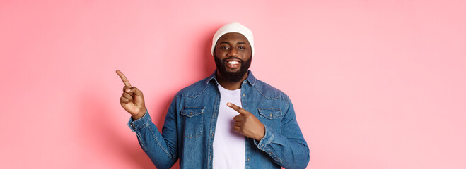 Happy african-american man smiling, pointing fingers right and showing promo, making announcement, standing over pink background