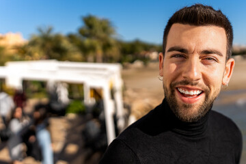Fototapeta na wymiar Positive young bearded male in black turtleneck with earrings looking at camera while standing on blurred background of tropical countryside area