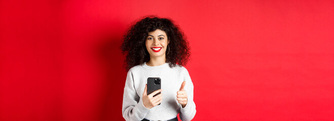 Smiling european woman looking happy and satisfied, using mobile phone and making thumb up,...