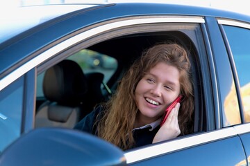 happy girl, young woman is sitting in her car, automobile, enjoying driving. Joyful lady in auto using, talking on cell mobile phone, calling on smartphone. Dangerous situation, careless driver