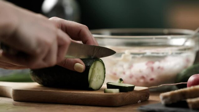 Close up of amateur cook cutting fresh cucumber into small chunks to prepare vegetable spread on wooden kitchen table; 4k 50fps