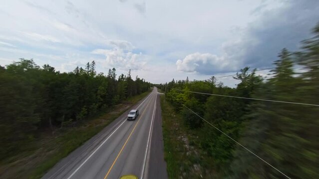 FPV drone shot flying over highway surrounded by woods while cars pass before gold sports car emerges at bottom of frame