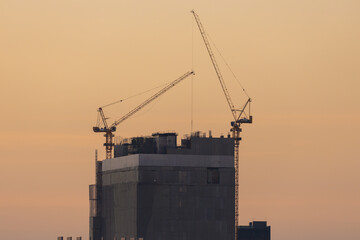 Aerial view of a large construction of a residential area with many multi-storey buildings under construction, a lot of tower cranes, workers, construction equipment at sunset