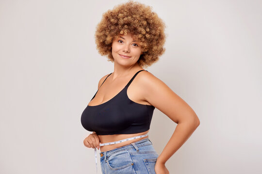 Positive curly haired caucasian woman with large breasts measures her parameters with a measuring tape, isolated next to white