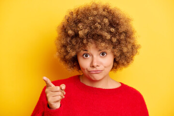 Woman pointing her index finger at the camera, choosing you, she is standing dressed in a red sweater with a funny expression, isolated on a yellow background