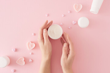 Women's cosmetics concept. Flat lay composition made of cream jar in hands, heart shaped candles, cosmetic bottles and heart baubles on pastel pink background. Mother's day cosmetic idea.