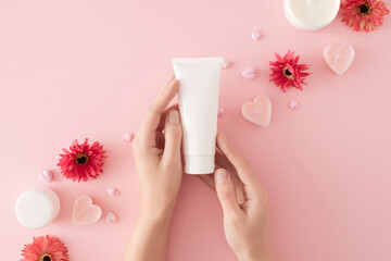 Women skin care concept. First person top view photo of female hands holding tube cream, heart shaped candles, cream jars and flowers on pastel pink background. Mother's day cosmetic idea.