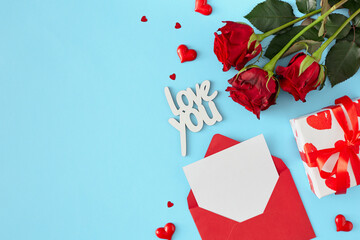 Women day concept. Top view photo of open envelope with white card, red flowers, gift box, hearts and inscription love you on blue background with copy space. 8-march holiday card idea.