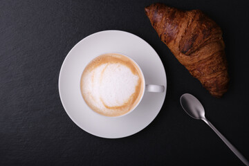 Cappuccino with croissant on a black background. White cup with cappuccino on a dark background flat lay. Italian breakfast on a black background.