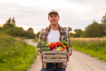 Farmer with a vegetable box in front of a sunset agricultural landscape. Man in a countryside field. Country life, food production, farming and country lifestyle concept.
