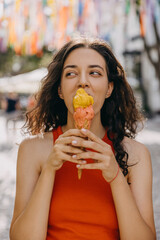 Happy young brunette woman on city street on a hot summer day, eating a big double layered ice...