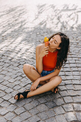 Young brunette woman sitting on city street on a hot summer day, eating an ice cream.