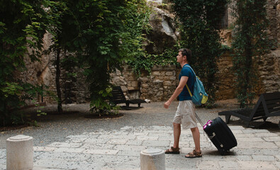 A tourist with a backpack and a suitcase walks along a stone wall with greenery. Travel and vacation