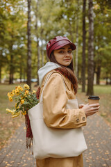 Side view of smiling young female with cup of coffee to go standing in park in fall season 