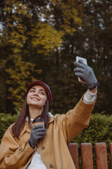 Delighted woman with coffee cup taking selfie in autumn park and having fun 