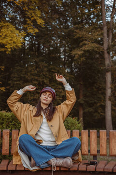Carefree woman with closed eyes enjoying music in earphones while sitting on bench in autumn park 