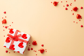 Saint Valentine's Day concept. Top view photo of white giftbox with red ribbon bow heart shaped candies and sprinkles on isolated light beige background with copyspace