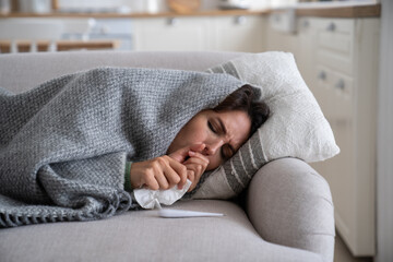 Young woman lying under blanket on sofa coughing and sneezing, using paper tissue to blow her nose. Unhealthy female caught cold or flu, resting on couch, suffering coronavirus. Cold homes and health