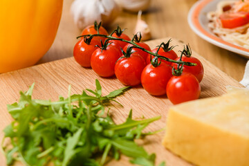 Sprig of cherry tomatoes on the table with garlic, parmesan cheese, arugula leaves and a bowl of pasta.