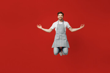 Full body young housewife housekeeper chef cook baker man wears grey apron jump high hold spread hands in yoga om aum gesture relax meditate try to calm down isolated on plain red background studio.