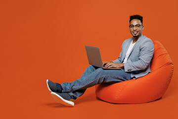 Full body young employee business man corporate lawyer wears formal grey suit shirt glasses work in office sit in bag chair hold use work on laptop pc computer isolated on plain red orange background.