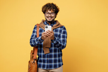 Young teen Indian boy IT student he wear casual clothes shirt glasses bag hold in hand use mobile cell phone chatting isolated on plain yellow color background High school university college concept.