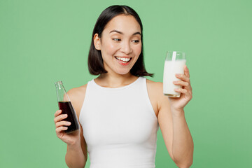 Young happy woman wear white clothes hold in hands bottle of soda pop fizzy cola glass of milk isolated on plain pastel green background. Proper nutrition healthy fast food unhealthy choice concept.