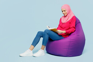 Full body young arabian muslim woman in pink abaya hijab sit in bag chair read book novel diary isolated on plain pastel light blue cyan background studio portrait People uae islam religious concept - obrazy, fototapety, plakaty