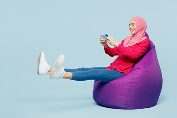Full body fun young arabian muslim woman wear pink abaya hijab sit in bag chair play racing app on...