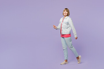 Full body side view happy fun elderly blonde woman 50s years old wear casual clothes denim jacket t-shirt walking going look camera isolated on plain pastel light purple background studio portrait.