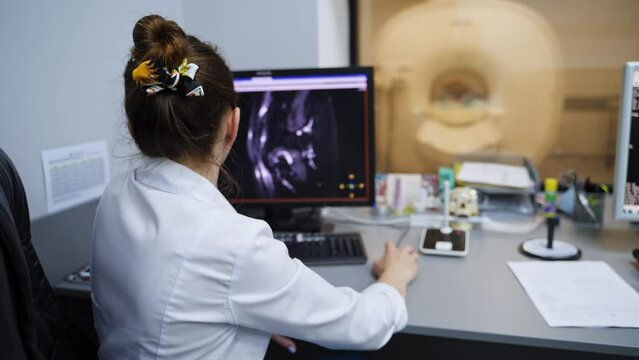 Rear view of a female lab technician sitting in front of computer. Expert looking through the diverse MRI scans on the screen.