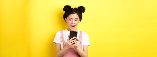 Pretty asian girl looking excited at screen, reading message on phone and smiling happy, standing...