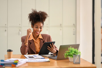 Happy African American businesswoman received good news, celebrating success with arms up in front of laptop at office desk