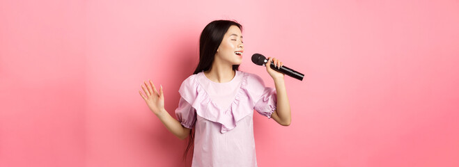 Beautiful asian girl perform song, singing in microphone and smiling romantic, standing in dress...