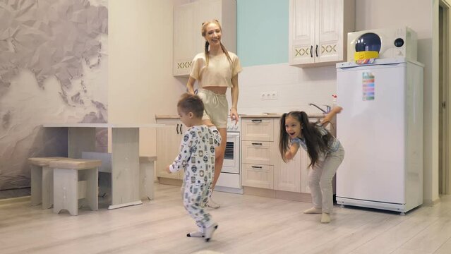 Young beautiful Caucasian girl is having fun and dancing with her little daughter and son on the same crutch in the kitchen of the house.Happy family having fun.Leg injury,bandage.Dancing on crutches