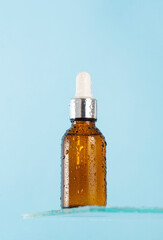 A brown dropper with a cosmetic product, with a serum for facial skin on a glass shelf with water splashes on blue background