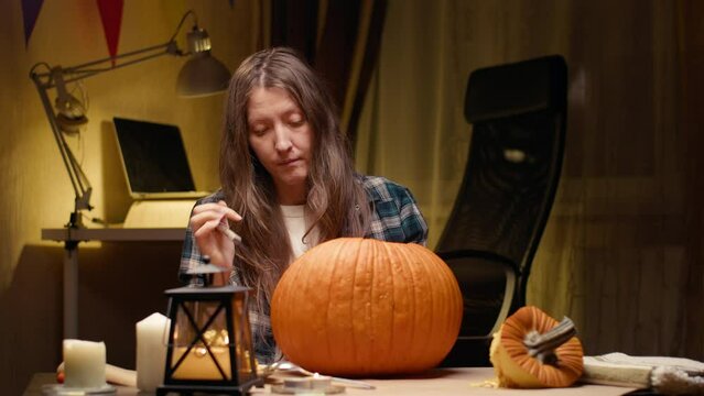 Preparing pumpkin for Halloween. Woman sitting and carving with knife face details of halloween Jack O Lantern pumpkin at home for her family.