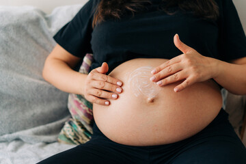 An unrecognizable pregnant woman carefully applies an anti-stretch mark cream to her belly skin.