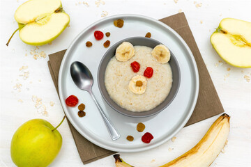 Funny cute kids childrens baby's healthy breakfast lunch oatmeal porridge in bowl look like bear face decorated with apple, banana, dried berry fruits. dessert food art on white wooden table